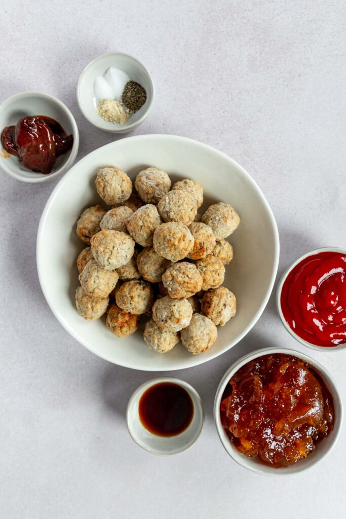 A bowl of chipotle peach meatballs is surrounded by small bowls containing ketchup, peach preserves, salt and pepper, jelly, and a dark liquid, all arranged on a light surface.