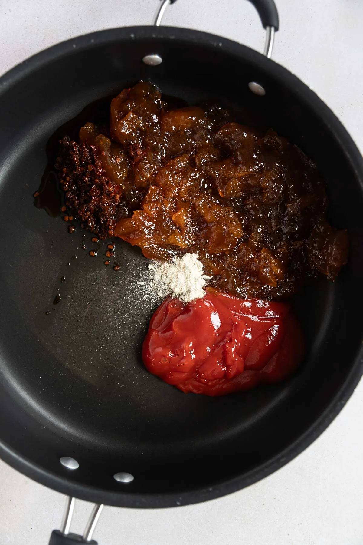 A black saucepan on a light surface contains spoonfuls of peach preserves, red ketchup, white powdered seasoning, and a dark paste—ingredients ready to be stirred together for delicious chipotle peach meatballs.