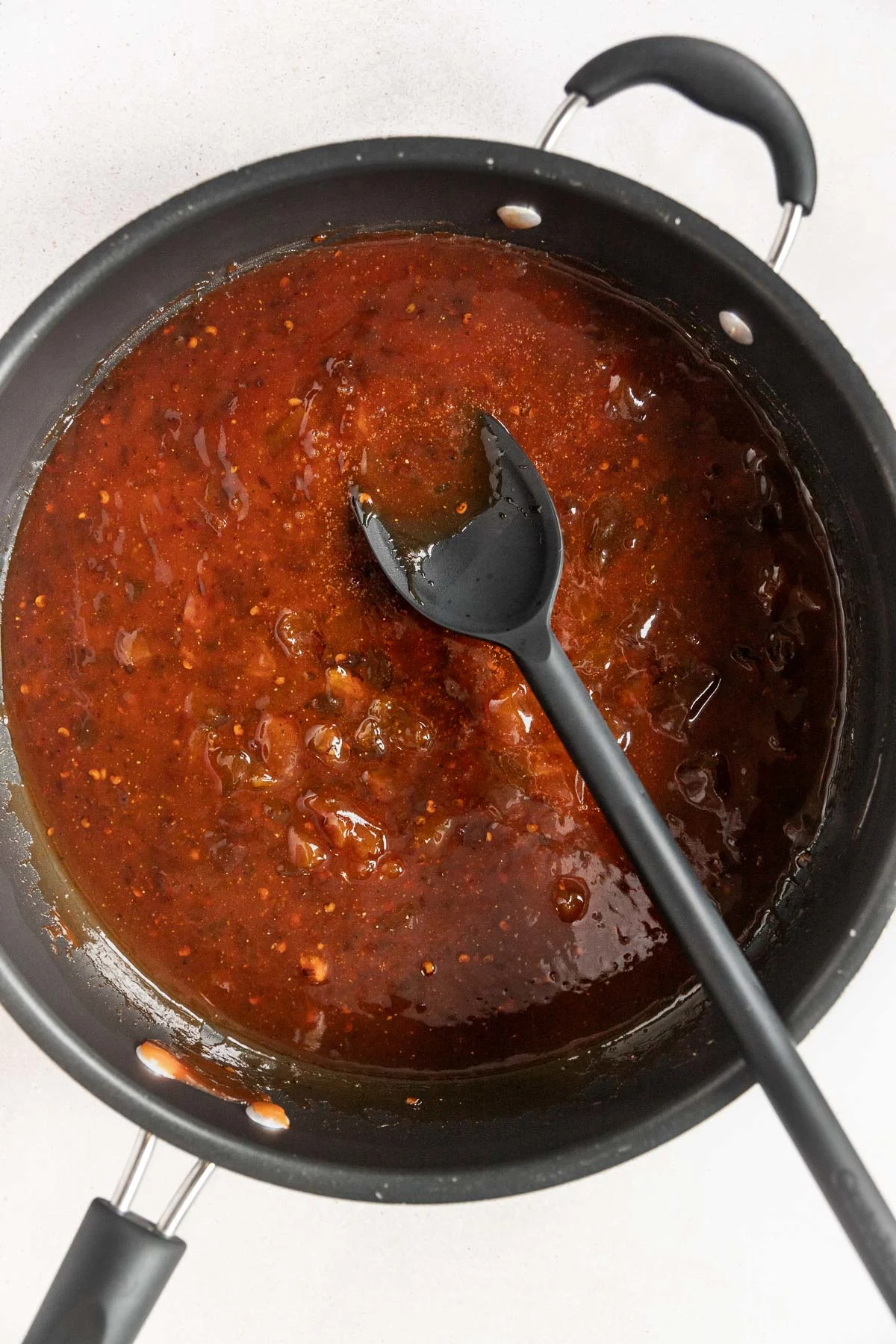 A black pan filled with thick, chunky red pasta sauce sits on a white surface. A black spoon rests inside, partially submerged in the sauce—perfect for serving alongside chipotle peach meatballs.