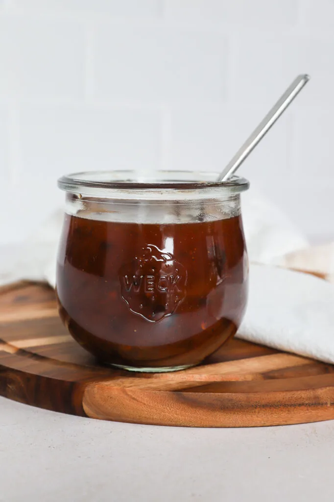 A glass jar filled with dark brown sauce for sweet and spicy meatballs sits on a round wooden board. A metal spoon rests inside the jar, and a white cloth is partially visible in the background.