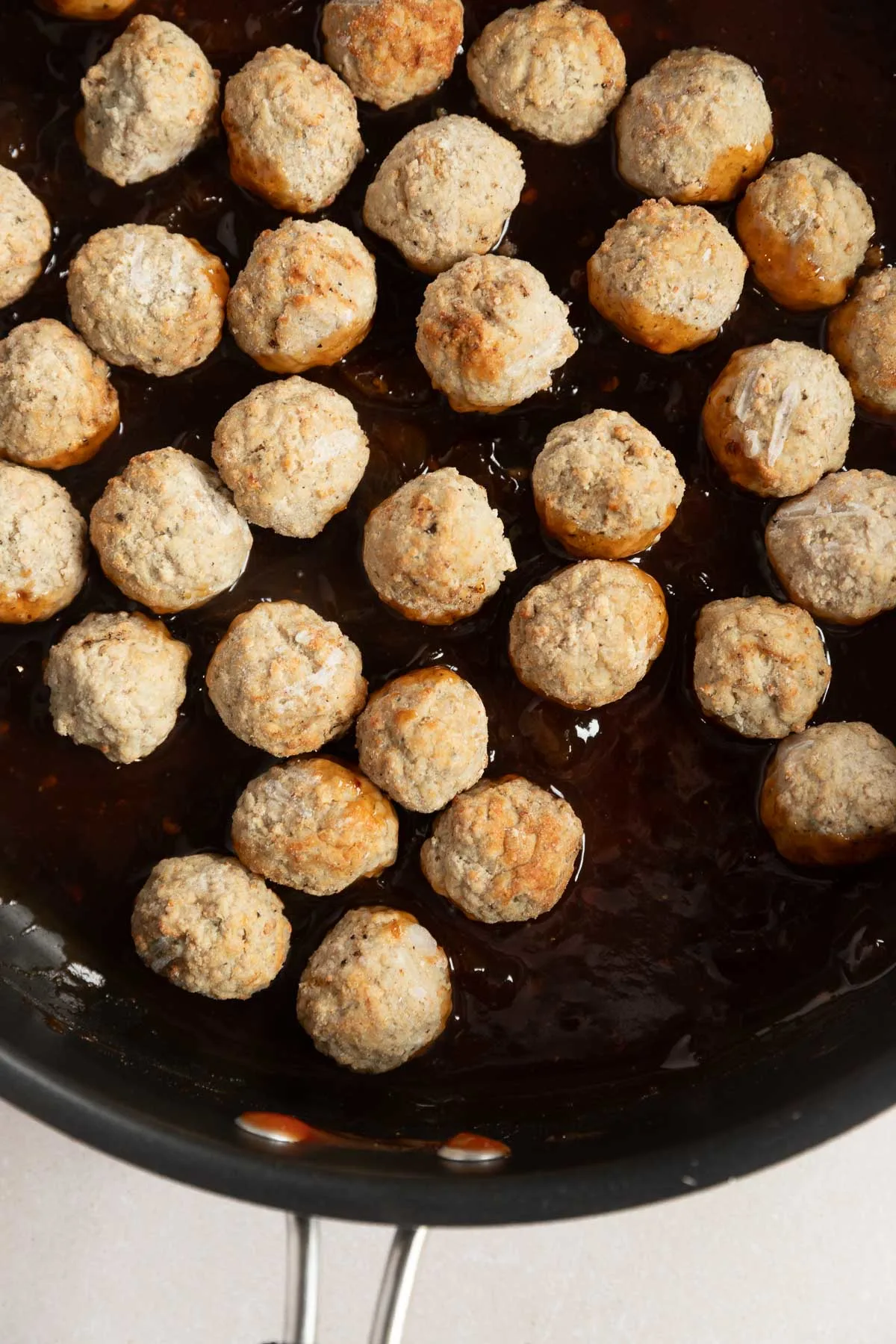 A pan filled with evenly spaced appetizer meatballs partially submerged in a dark, glossy sauce. The light brown meatballs sit invitingly in the pan, which features sturdy metal handles.