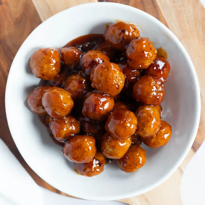 A white bowl filled with appetizer meatballs sits on a round wooden cutting board, viewed from above. The meatballs are coated in a shiny brown sauce.