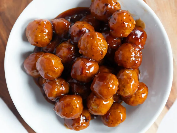 A white bowl filled with appetizer meatballs sits on a round wooden cutting board, viewed from above. The meatballs are coated in a shiny brown sauce.