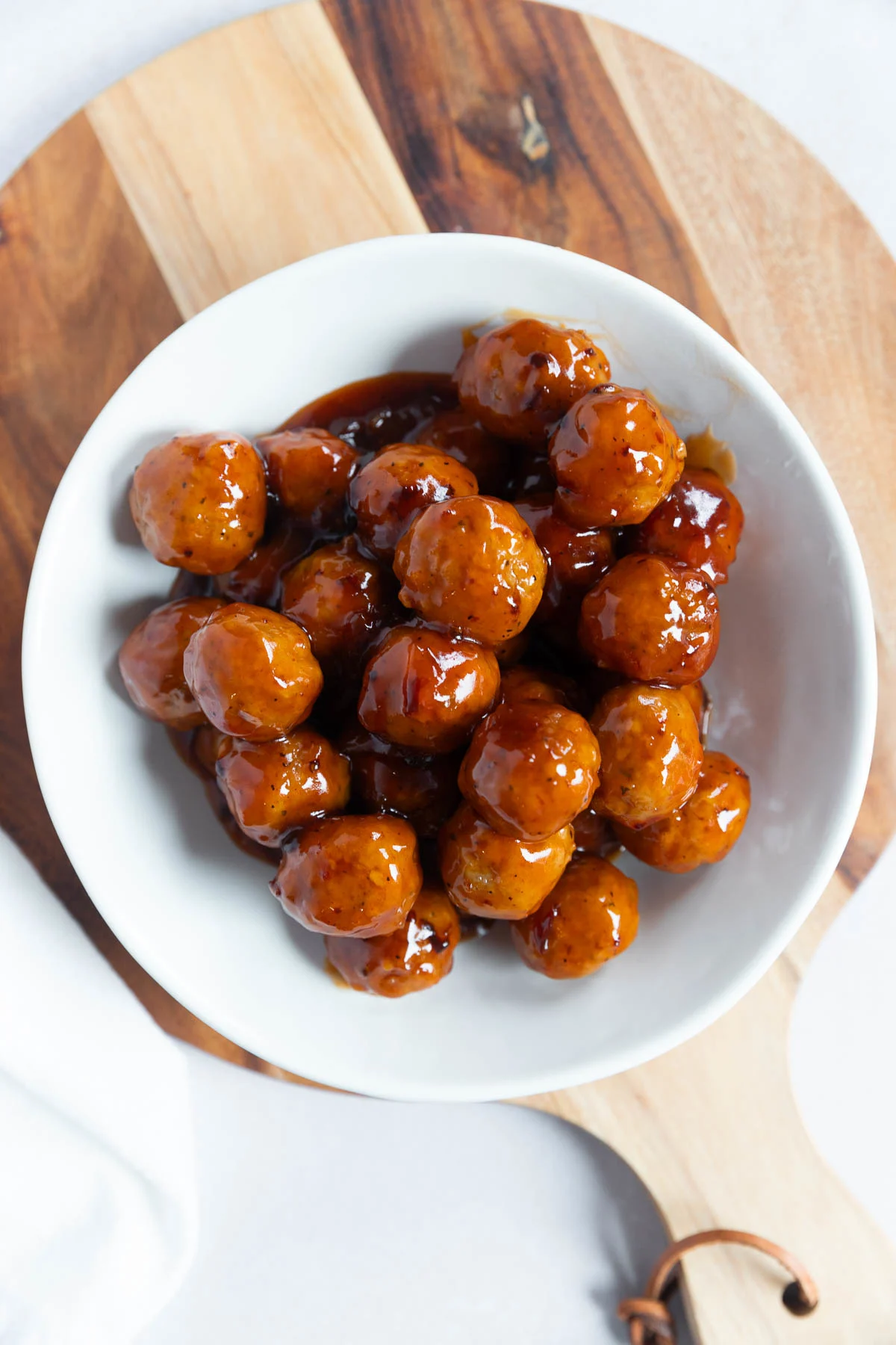 A white bowl filled with appetizer meatballs sits on a round wooden cutting board, viewed from above. The meatballs are coated in a shiny brown sauce.