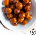 A white bowl filled with glazed chipotle peach appetizer meatballs sits on a wooden surface. Text at the bottom reads, Chipotle Peach Meatballs, The Culinary Compass.