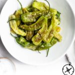 A white bowl filled with easy shishito peppers, drizzled with oil and sprinkled with salt, sits on a white surface. Text below the bowl reads, How to Cook Shishito Peppers, The Culinary Compass.