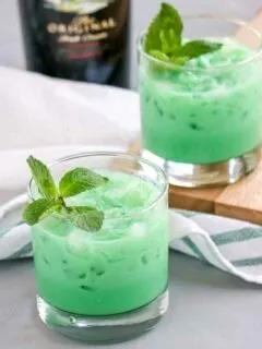 Two glasses of a minty green cocktail are placed on a wooden board. Each drink is garnished with fresh mint leaves. A bottle and striped cloth are in the background on a gray surface.