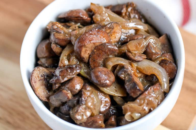 A white bowl filled with caramelized mushrooms and onions in a rich brown sauce, placed on a wooden surface.