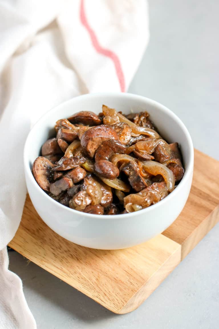 A white bowl filled with cooked chicken livers, caramelized onions and mushrooms sits on a wooden board, with a white cloth nearby.