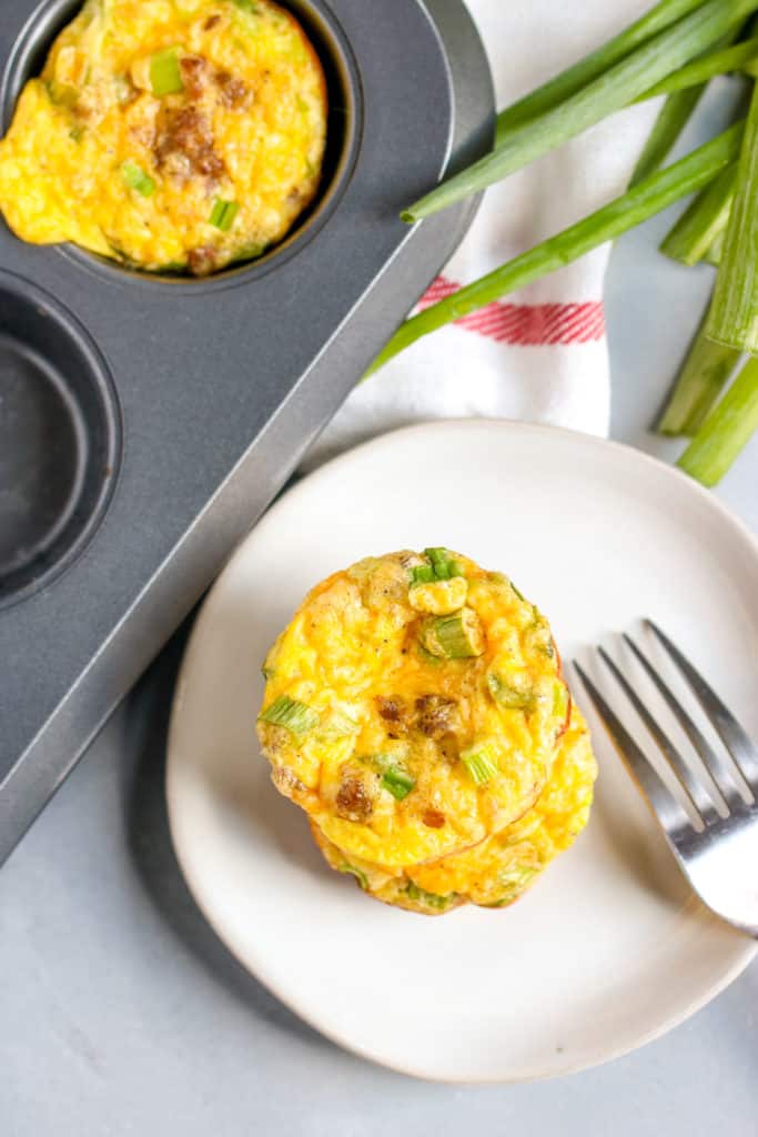 two sausage egg muffins stacked on top of a plate next to the muffin tin with green onions