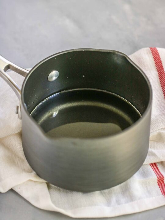 A small metal saucepan filled with clear liquid, perfect for learning how to make simple syrup, sits on a white cloth with a red stripe, placed on a light gray surface.