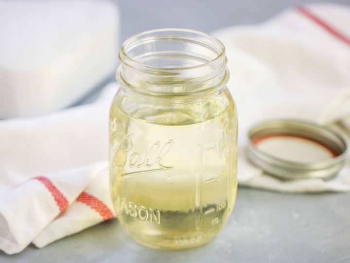 A glass mason jar filled with a light yellow simple syrup for cocktails sits on a gray surface next to a white towel with a red stripe and a metal jar lid.
