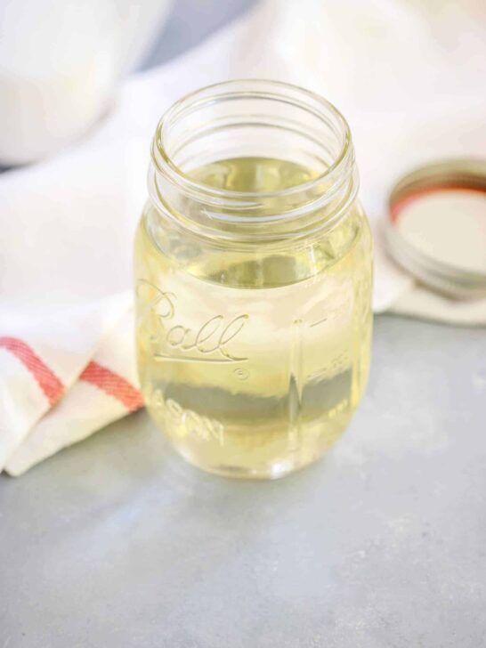 A clear mason jar filled with a light yellow simple syrup sits on a gray surface next to a white cloth with a red stripe and a mason jar lid, ready for your next simple syrup recipe.
