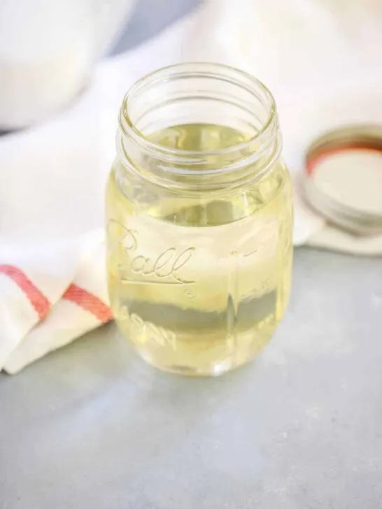 A clear mason jar filled with a light yellow simple syrup sits on a gray surface next to a white cloth with a red stripe and a mason jar lid, ready for your next simple syrup recipe.