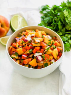 a close up of peach salsa in a bowl with a peach, lime, and cilantro next to it