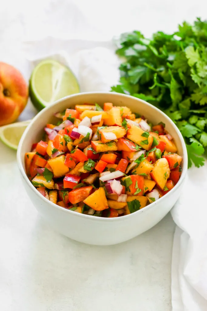 a close up of peach salsa in a bowl with a peach, lime, and cilantro next to it