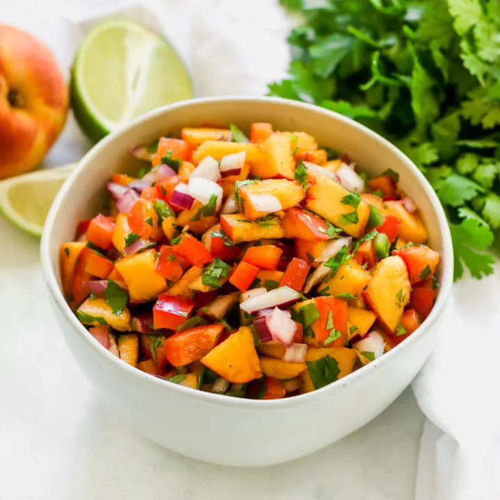 a close up of peach salsa in a bowl with a peach, lime, and cilantro next to it