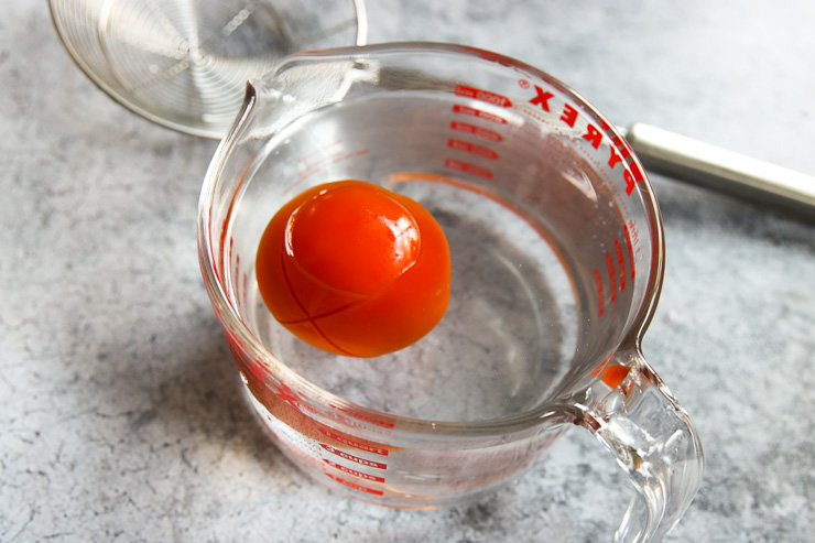 a scored tomato floating in water in a pyrex measuring cup