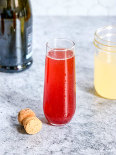 A glass of Pomegranate Mimosa next to a champagne bottle, cork and mason jar of Sour Mix