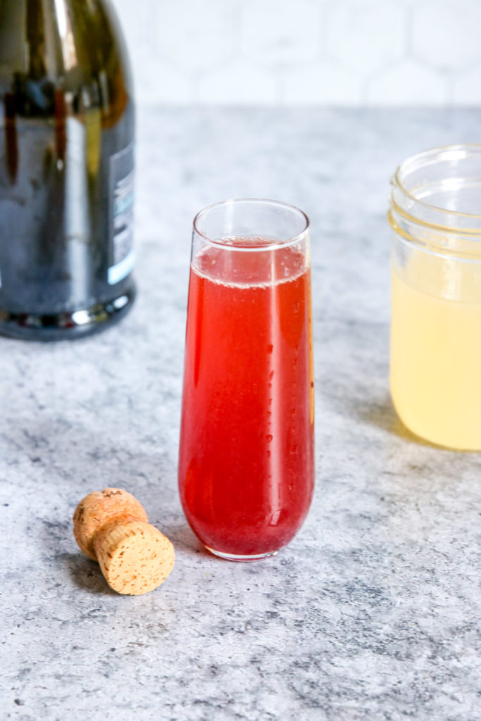 A glass of Pomegranate Mimosa next to a champagne bottle, cork and mason jar of Sour Mix