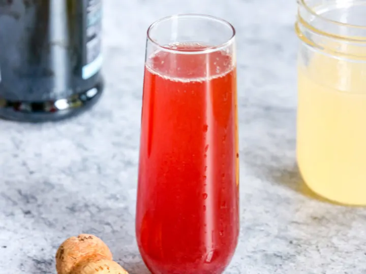 A glass of Pomegranate Mimosa next to a champagne bottle, cork and mason jar of Sour Mix