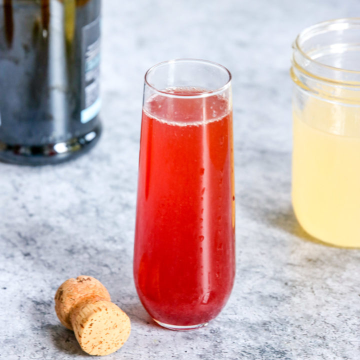 A glass of Pomegranate Mimosa next to a champagne bottle, cork and mason jar of Sour Mix