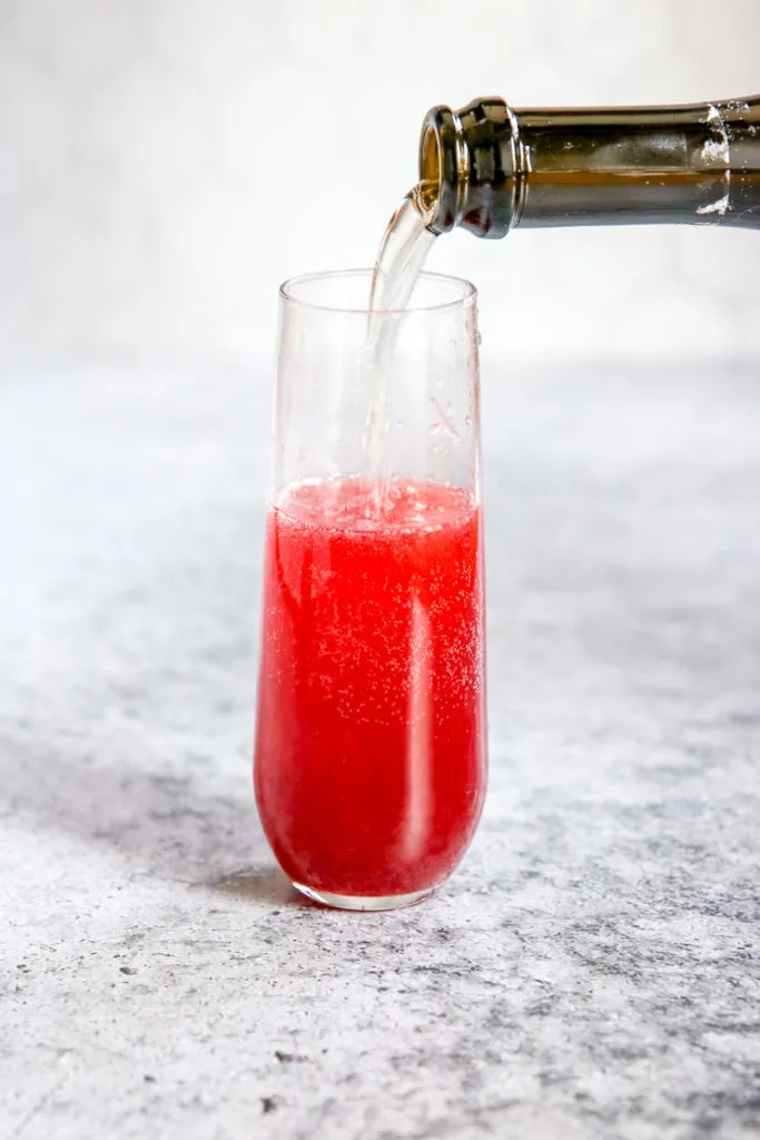 A side view of a bottle pouring champagne into a glass filled with Pomegranate Mimosa