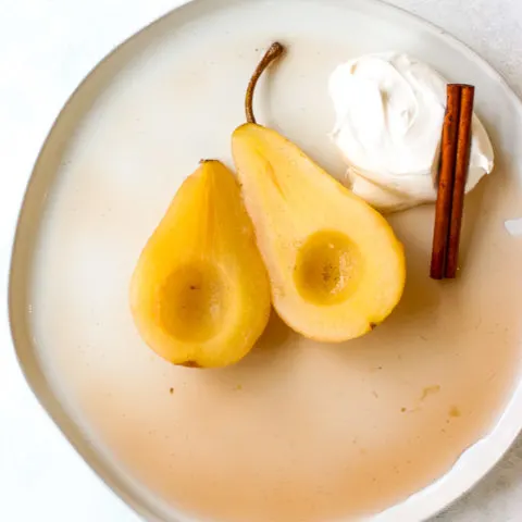 a plate of two halves of poached pears with whipped cream and a cinnamon stick