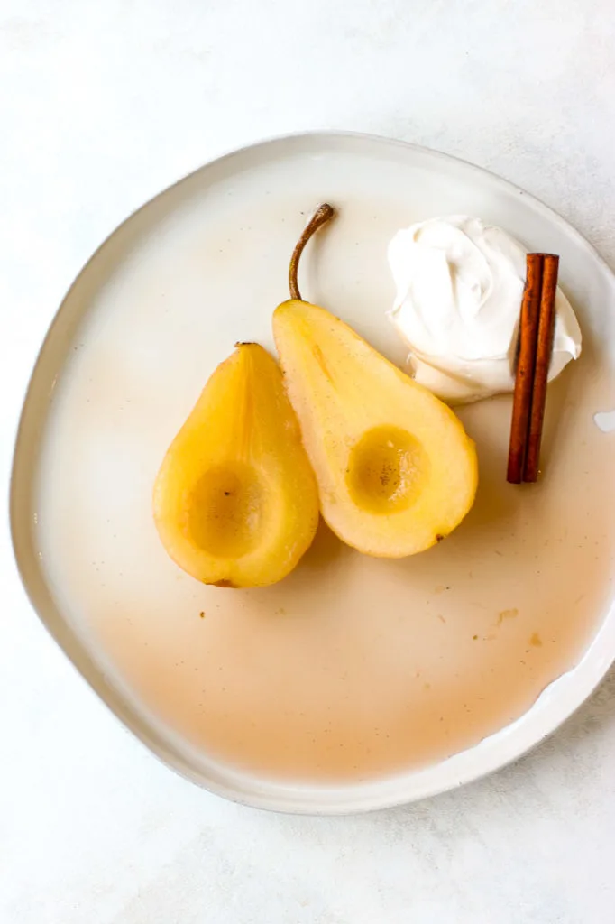 a plate of two halves of poached pears with whipped cream and a cinnamon stick