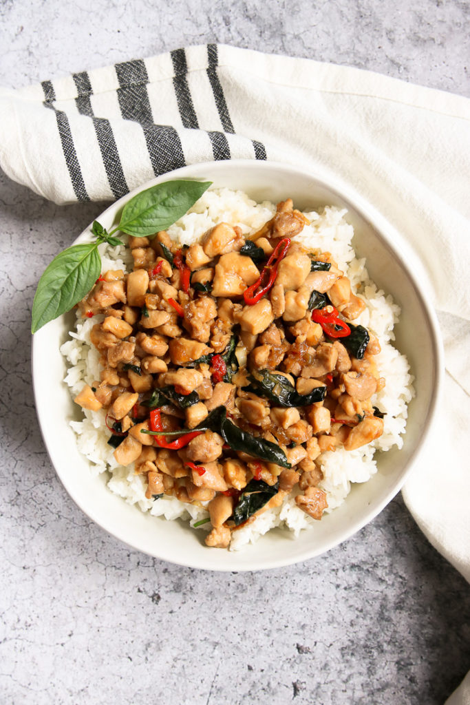 an overhead shot of a bowl of thai basil chicken over rice with a sprig of thai basil