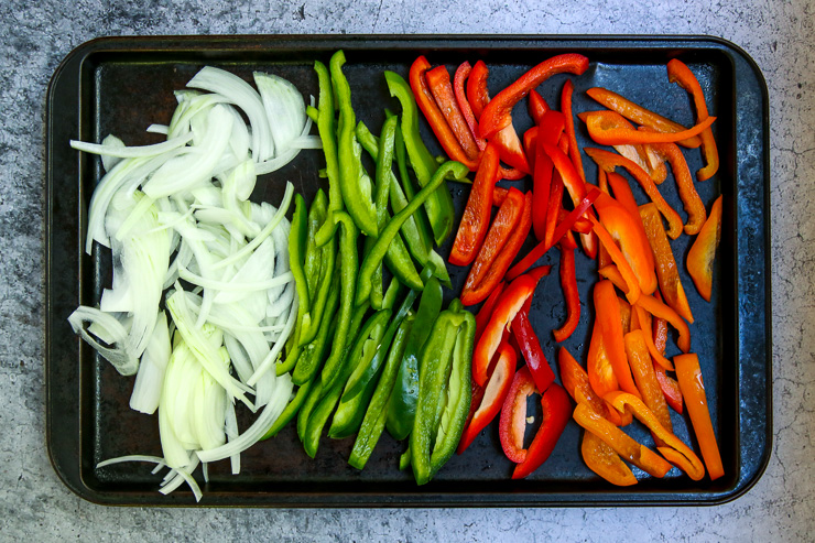 a cookie sheet full of raw, sliced onion, green bell peppers, red bell peppers, and orange bell peppers