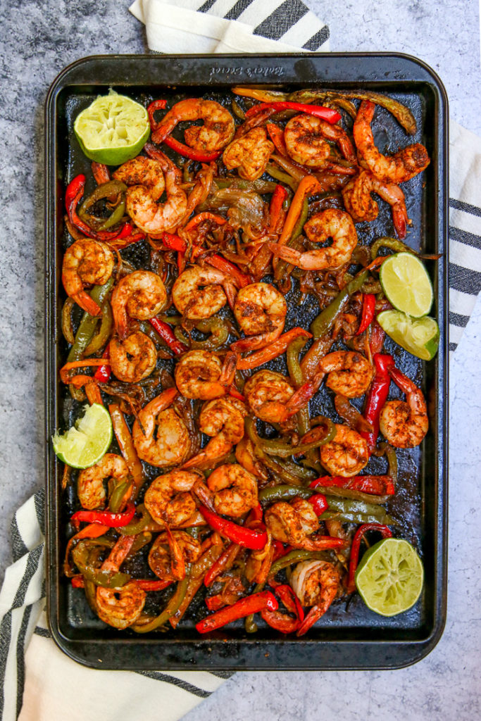 a cookie sheet full of cooked shrimp, onions, and peppers with lime wedges