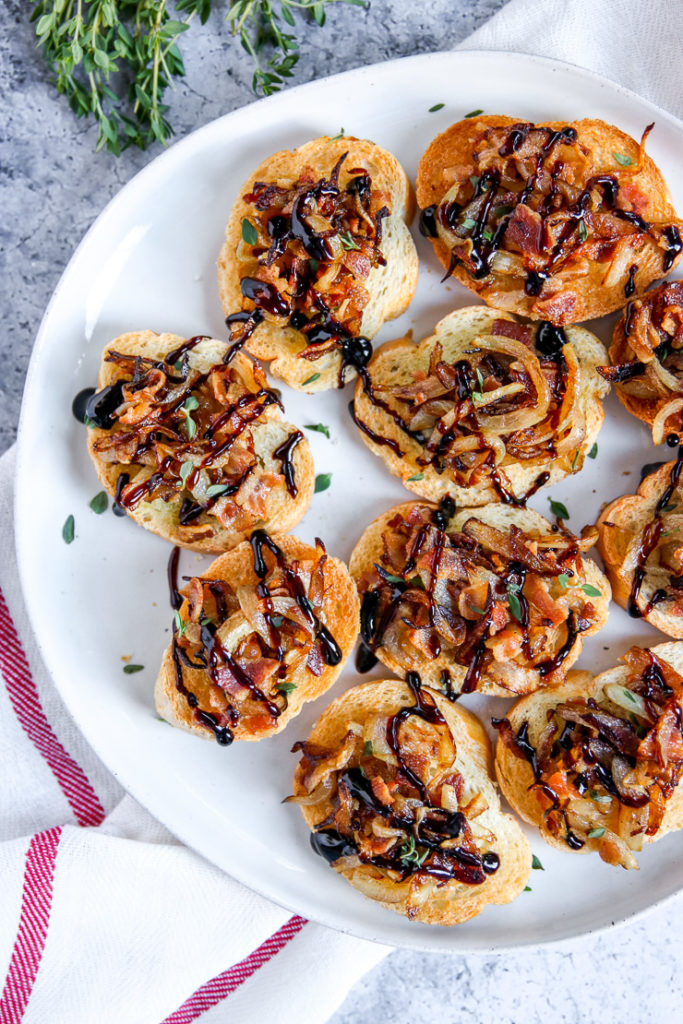 a few pieces of crostini on a plate topped with bacon, caramelized onion, thyme, and balsamic glaze