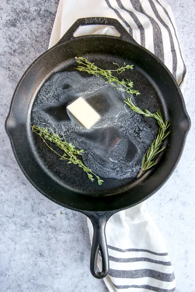 a cast iron skillet with partially melted butter, thyme, and rosemary