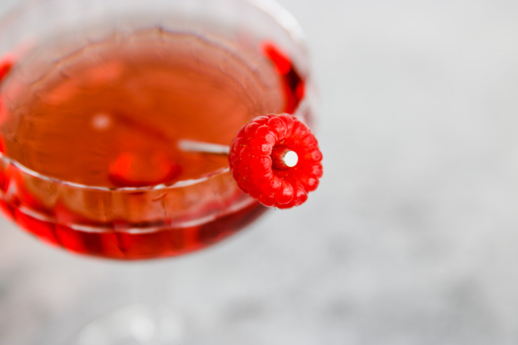 a close up of a raspberry on a metal cocktail pick sitting in a coupe glass