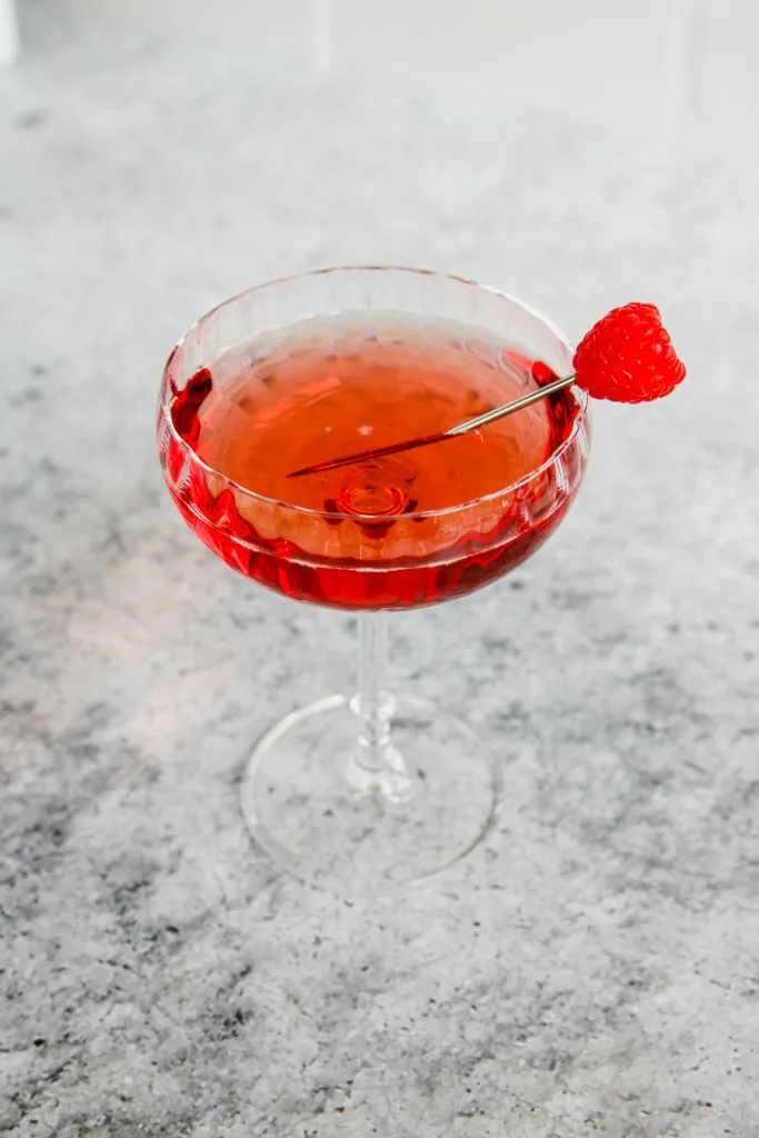 a coupe glass of chambord and champagne with a raspberry on a metal cocktail pick