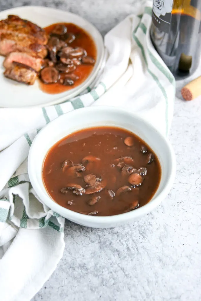 a close up of a white bowl of red wine mushrooms with a bottle of red wine and steak in the background