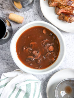 a bowl of red wine mushrooms next to a cut steak, wine bottle, glass, and cork