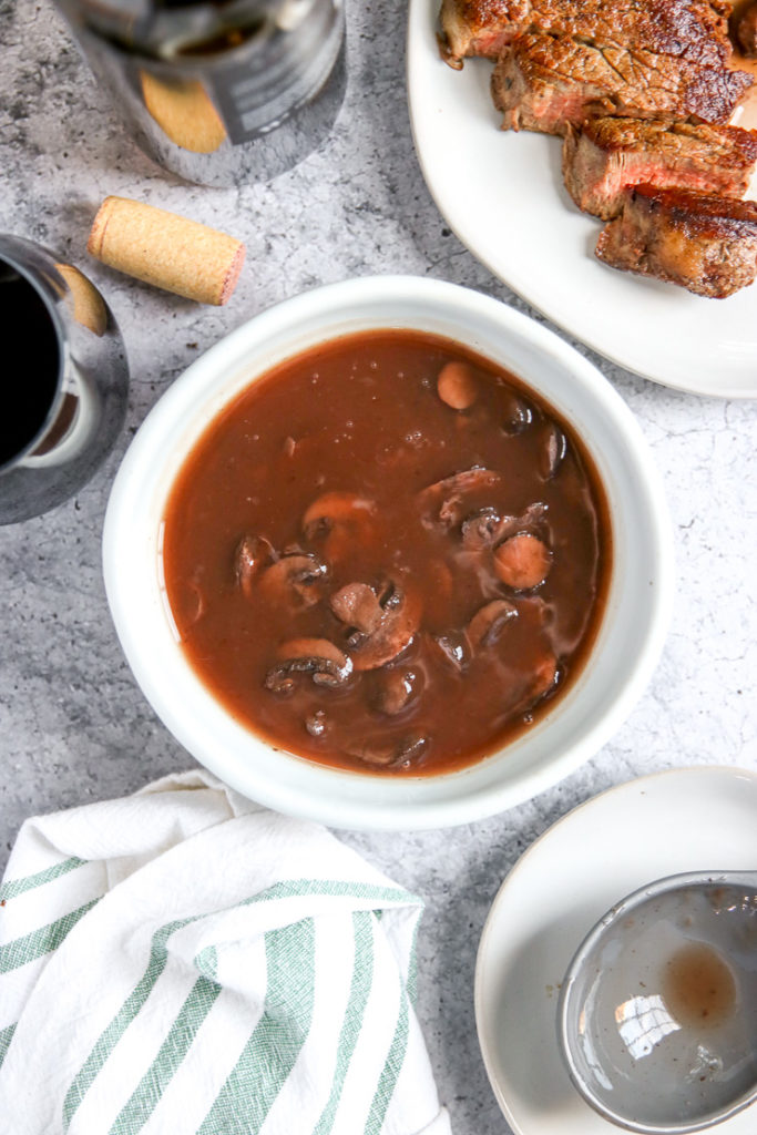 a bowl of red wine mushrooms next to a cut steak, wine bottle, glass, and cork