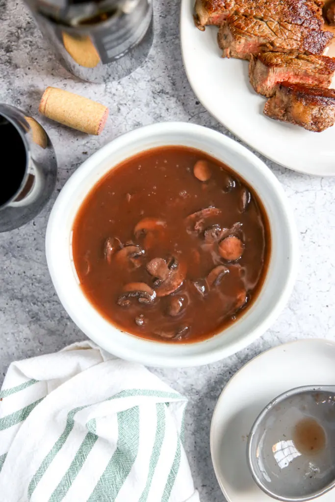 a bowl of red wine mushrooms next to a cut steak, wine bottle, glass, and cork