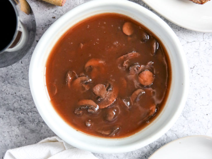 a bowl of red wine mushrooms next to a cut steak, wine bottle, glass, and cork