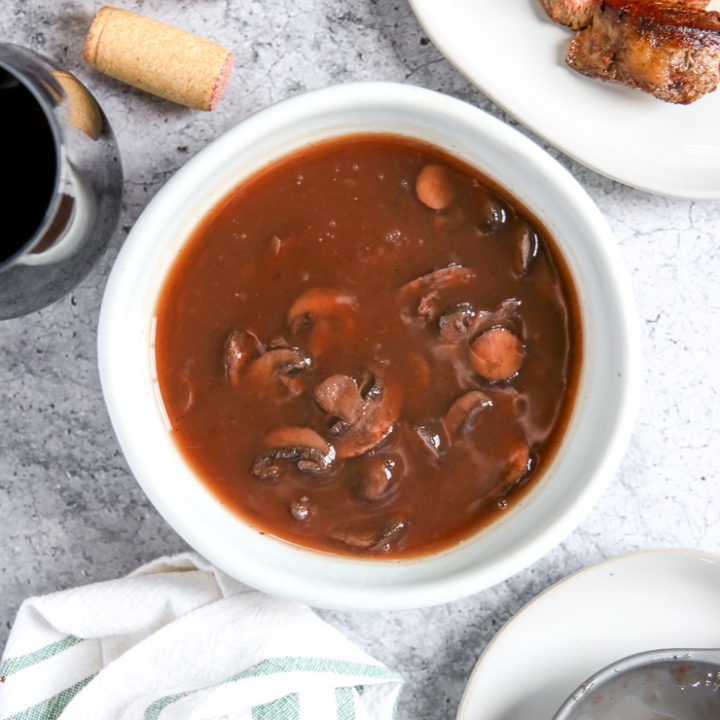 a bowl of red wine mushrooms next to a cut steak, wine bottle, glass, and cork