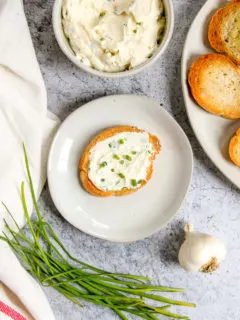 an overhead shot of a slice of crostini with roasted garlic spread on top next to garlic and chives