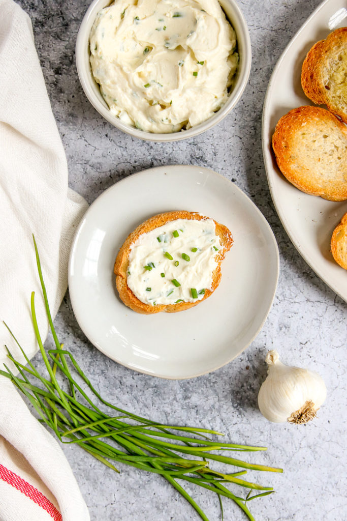 an overhead shot of a slice of crostini with roasted garlic spread on top next to garlic and chives