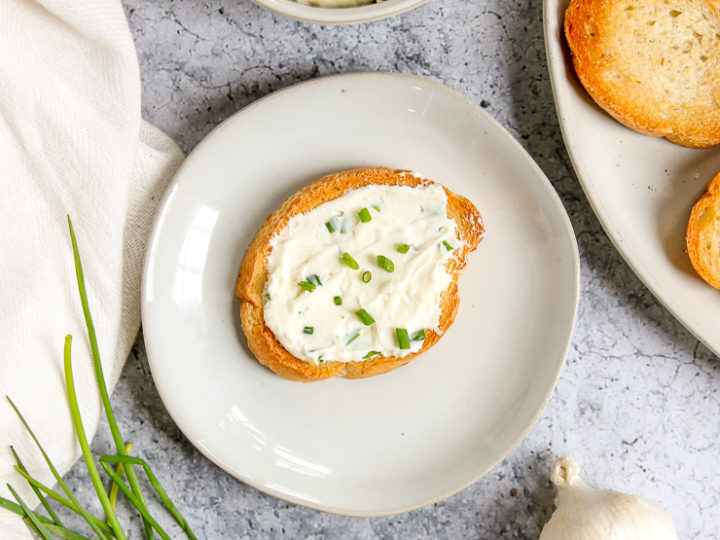 an overhead shot of a slice of crostini with roasted garlic spread on top next to garlic and chives