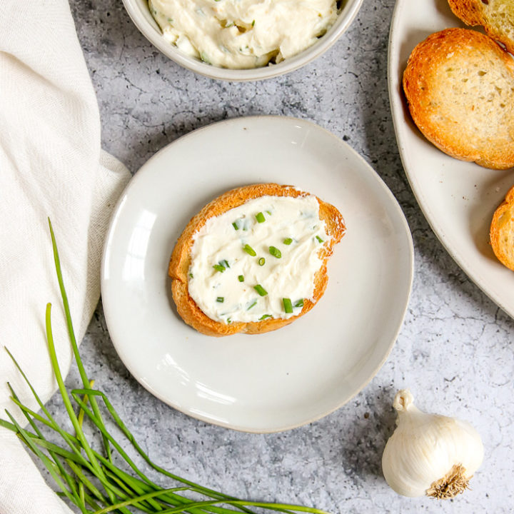 an overhead shot of a slice of crostini with roasted garlic spread on top next to garlic and chives