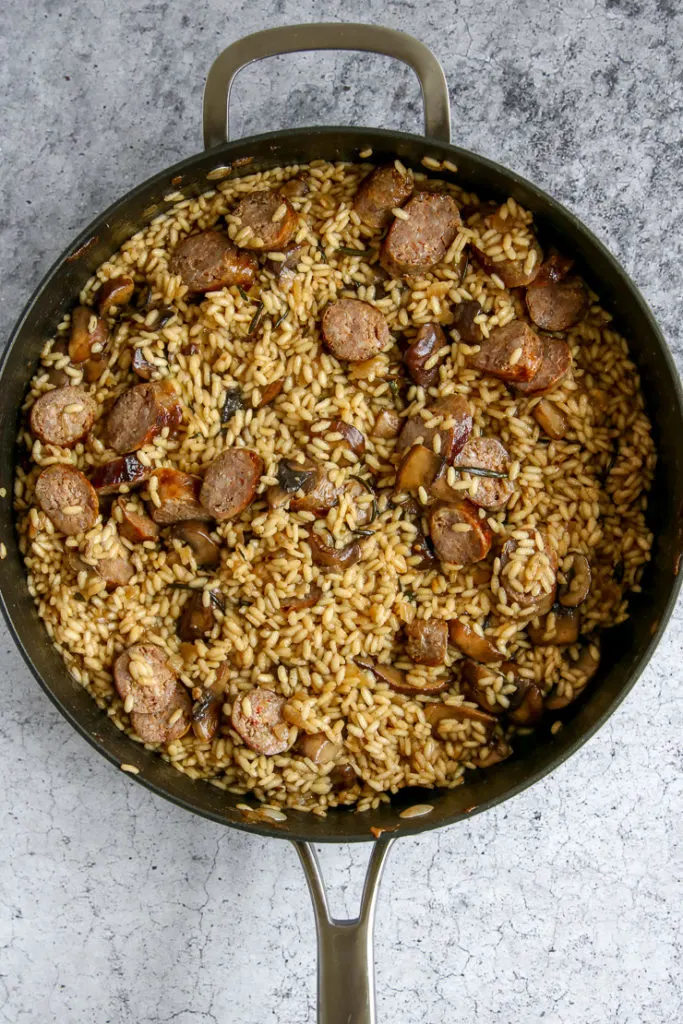 a large skillet full of sliced sausage, herbs, sliced mushrooms, and arborio rice