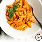A white bowl filled with penne pasta coated in creamy orange vodka sauce, garnished with fresh basil leaves and grated cheese, with a fork resting in the bowl. Text at the bottom reads Vodka Sauce - The Culinary Compass.