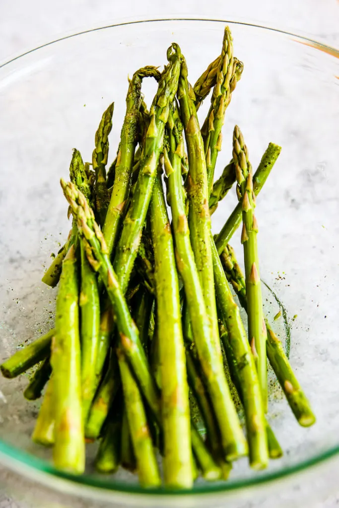 a glass bowl of asparagus stalks with olive oil, salt and pepper on them