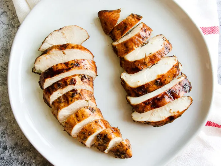 two sliced balsamic grilled chicken breasts on a white plate sitting on a white and red striped napkin